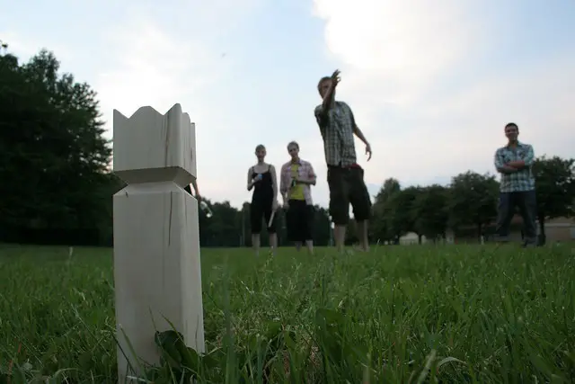 view from the grass looking up at man throwing wooden baton, wooden Kubb king piece in the foreground