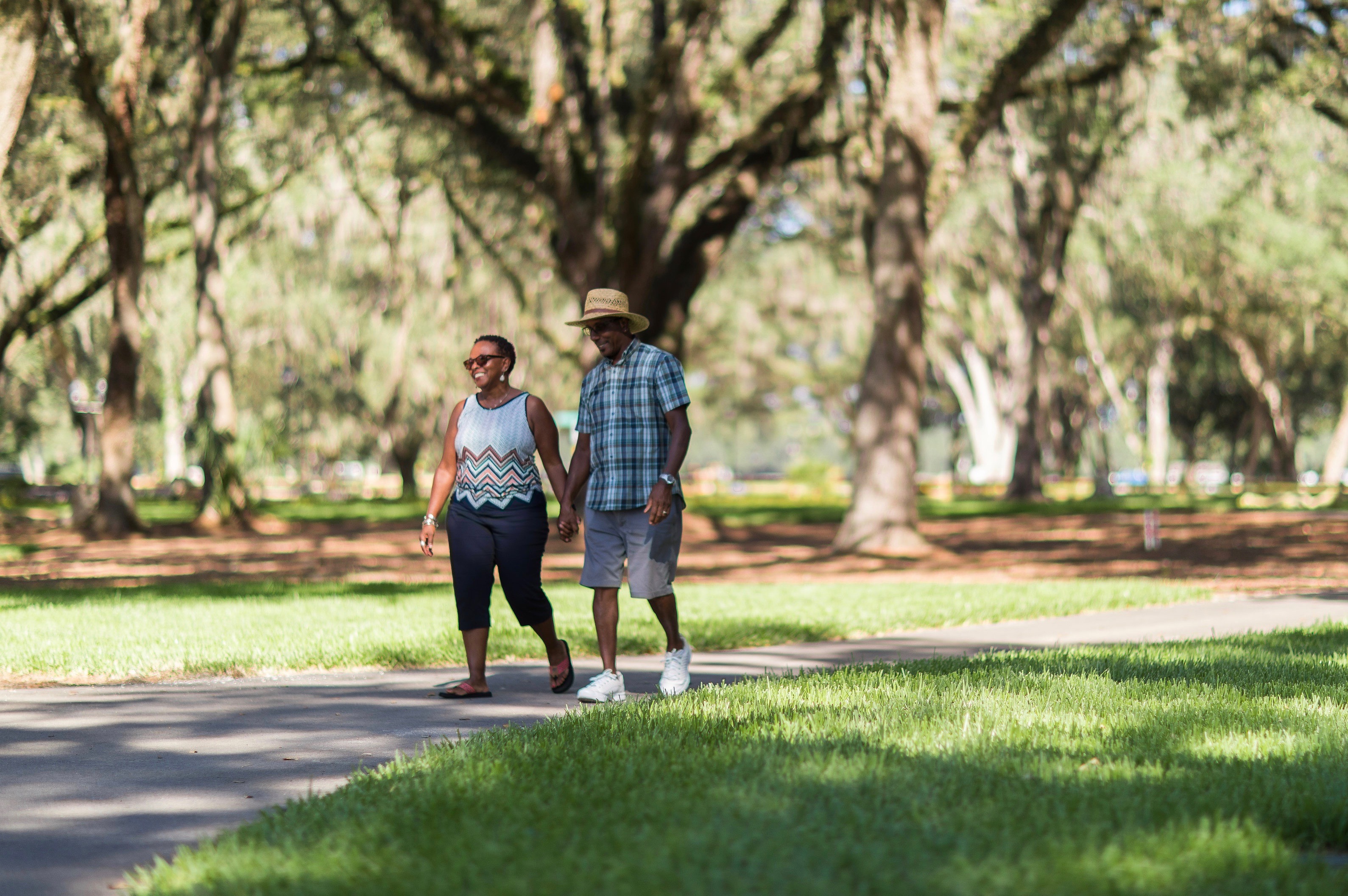 couple holding hands, smiling, walking on a paved path in sunny park with green grass on either side of path. Trees blurred in background