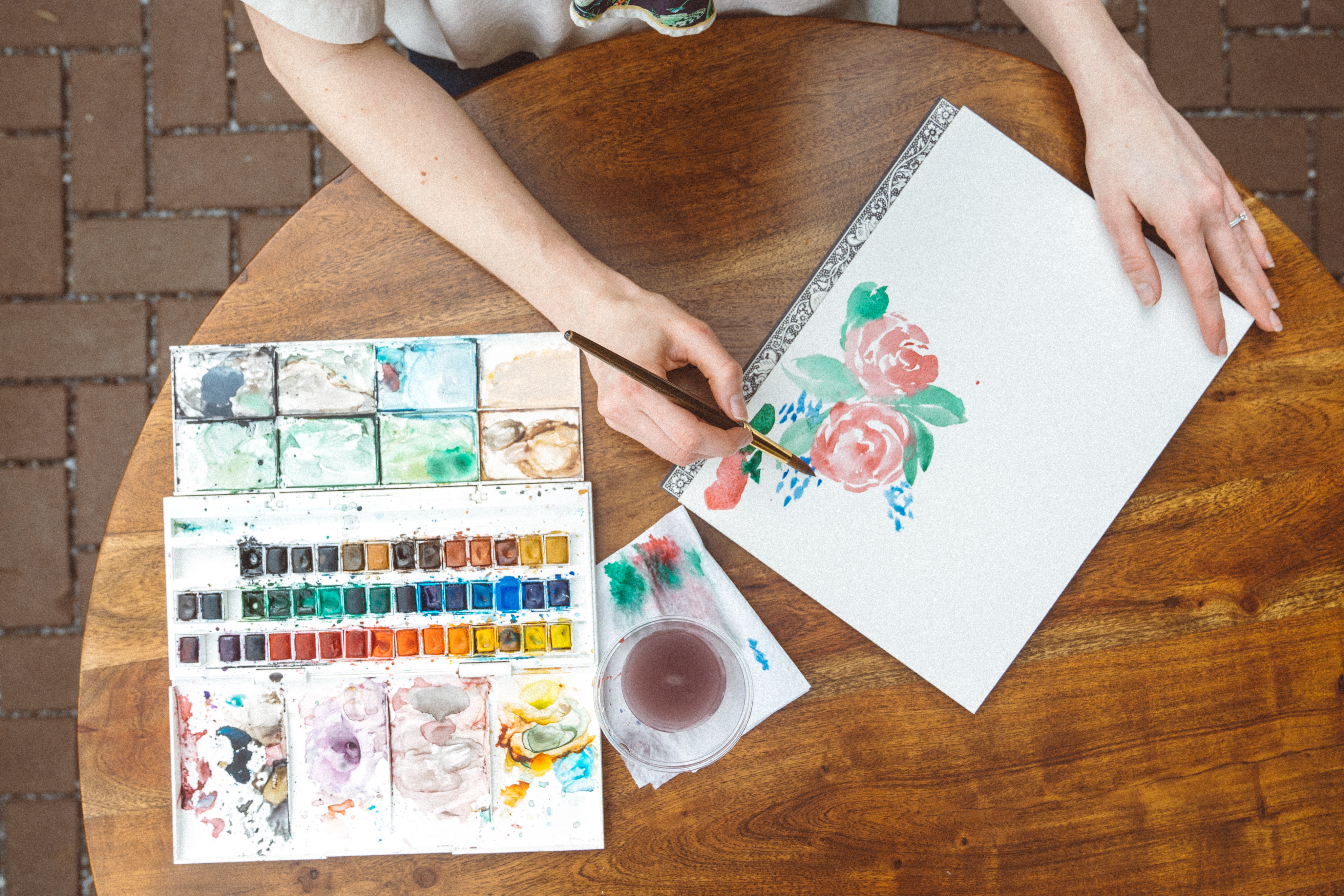 overhead view of woman painting red flower with watercolor paints. Open watercolor paint set on round wooden table beside her