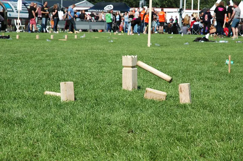 grass field with Kubb pieces displaying game play. Wooden baton midair about to strike wooden king piece