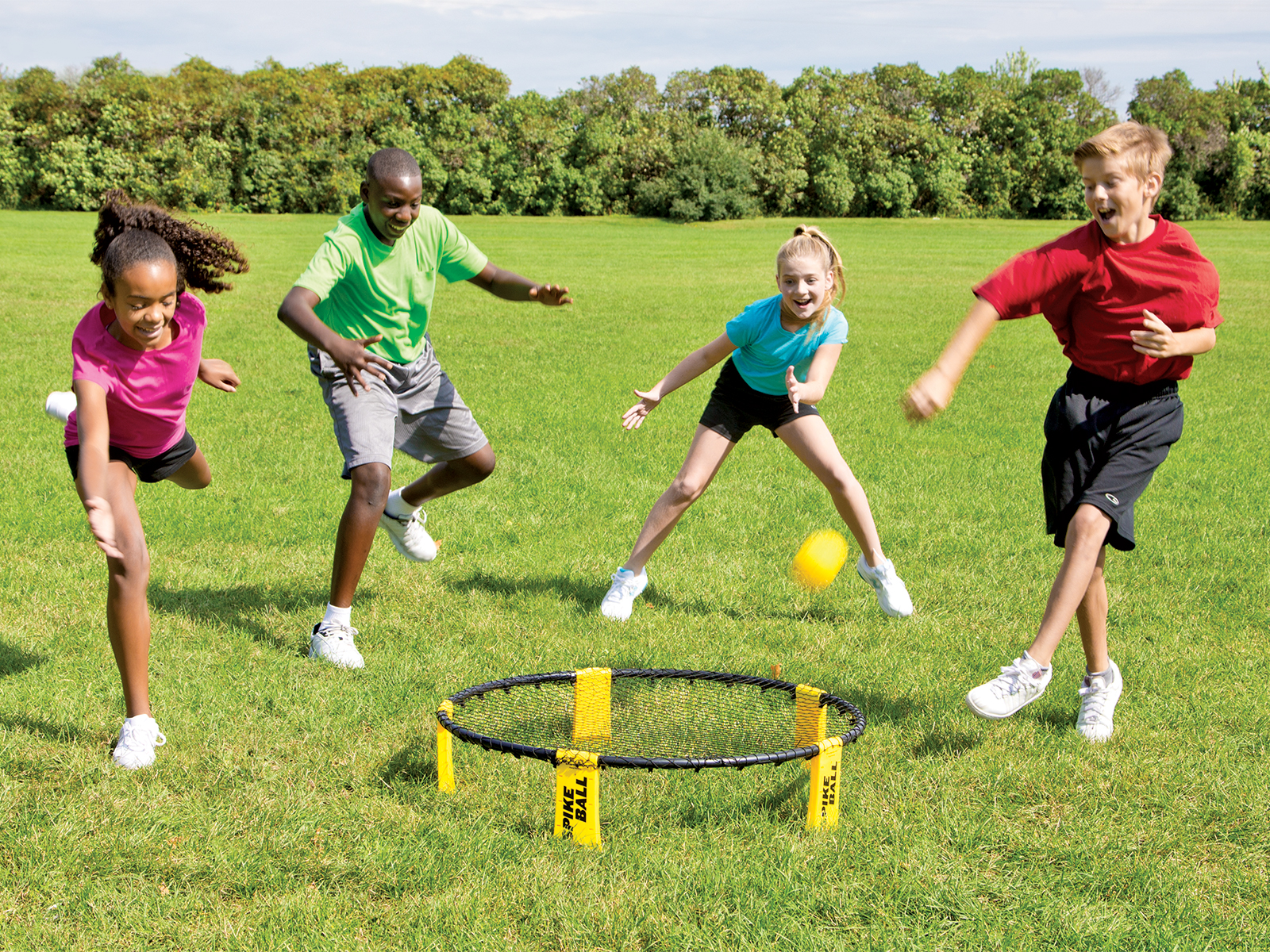 4 children ages 10-13 smiling and standing in a semi-circle around a small net on the group. Children are watching a ball bouncing off of the net