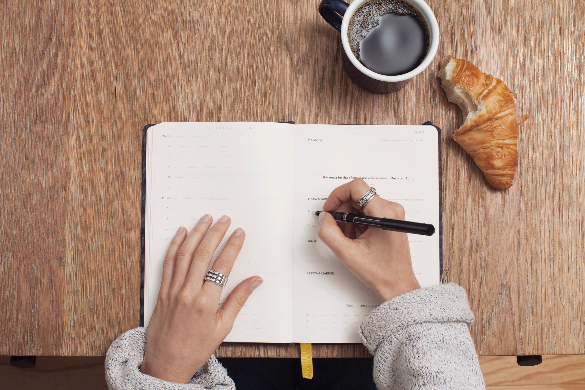 overhead view of woman journaling on top of wooden table with croissant and black coffee 