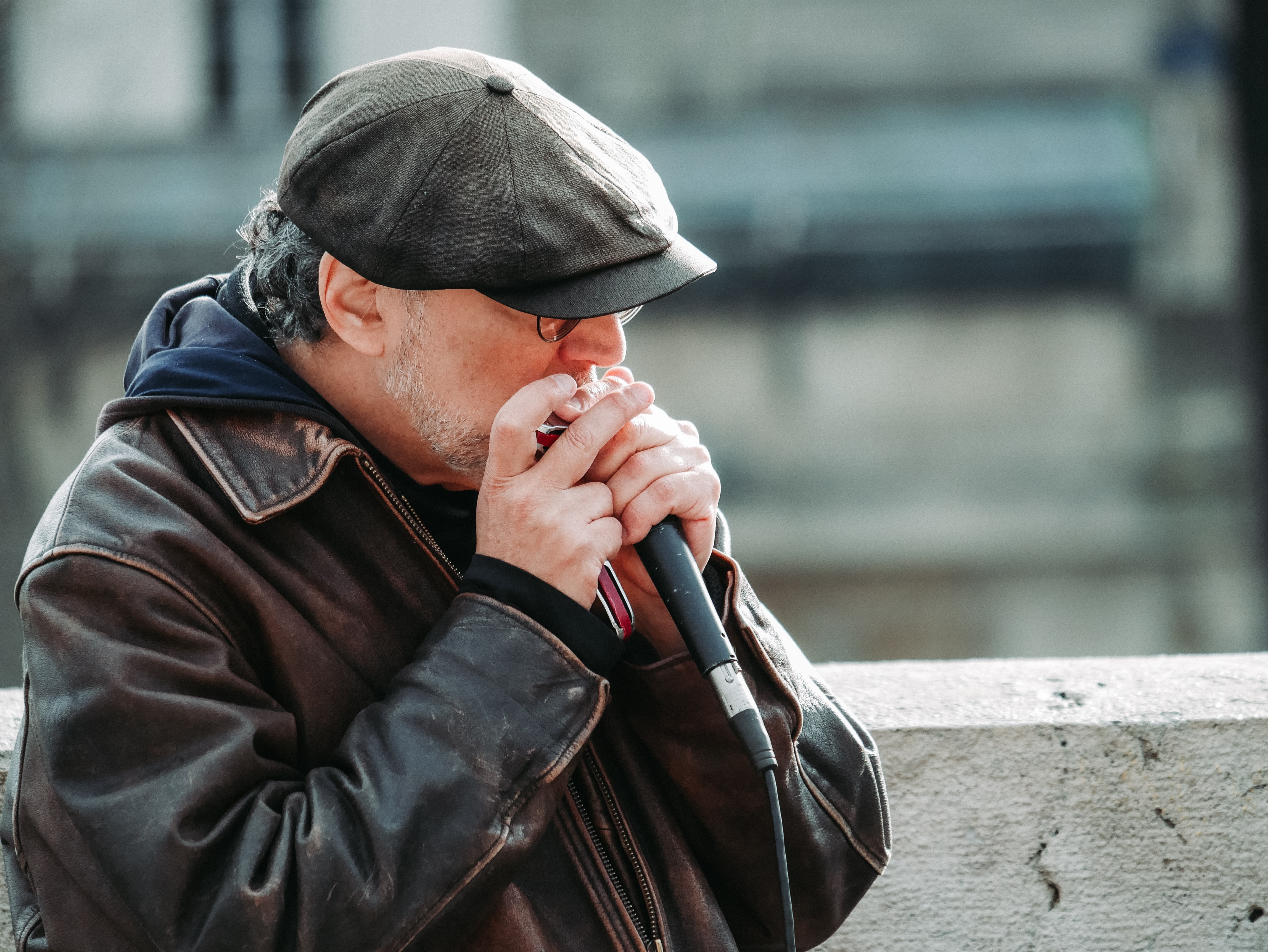 sixtyish year old man outdoors playing harmonica into a microphone. Wearing worn leather jacket and newspaper hat. Short grey beard and glasses.