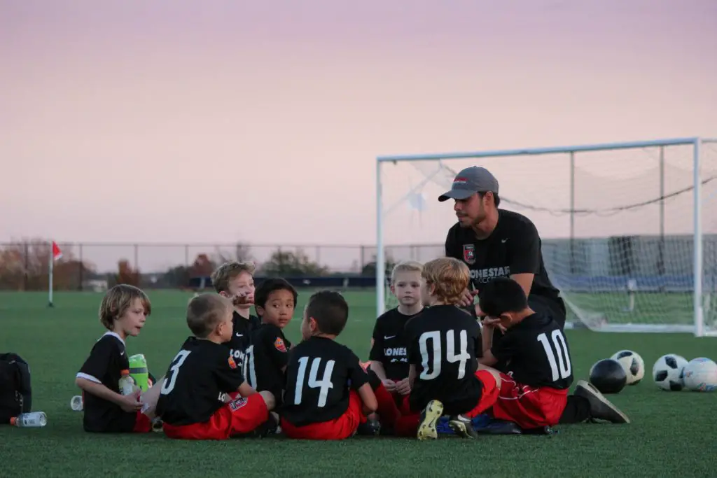 young adult man coaching boys soccer team; hudled on field talking sunset in the background