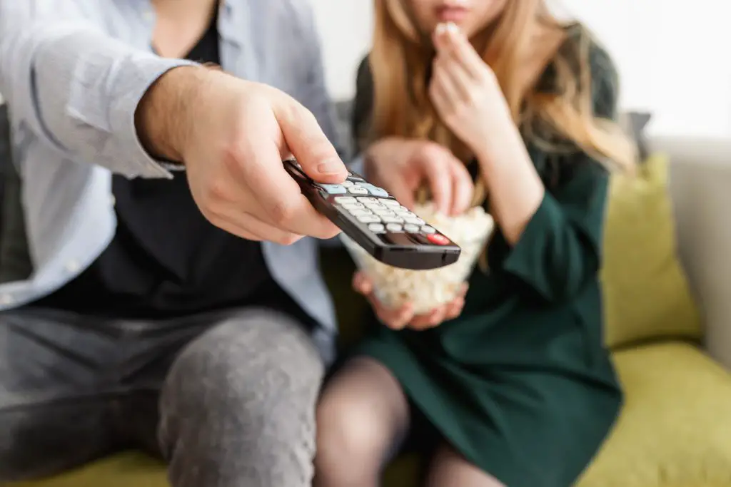 mans hand holding remote control, woman in the background eating popcorn