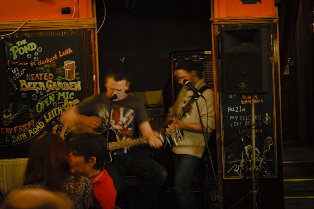 two men playing guitar in front of small crowd at open mic night