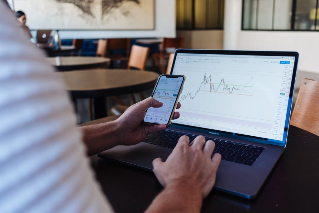 Man holding a cellphone and looking at an open laptop.  both devices open to stock charts. Hobbies for stay-at-home dads include stock market investing