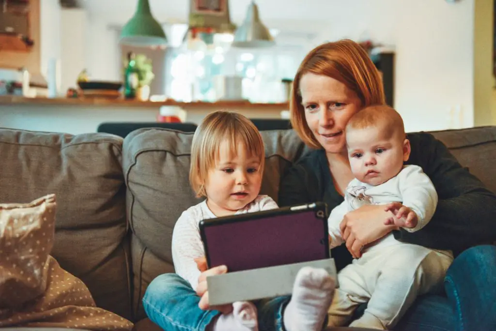 mother with toddler and baby sitting on a couch looking at a tablet.  Hobby for mothers and daughters playing video games