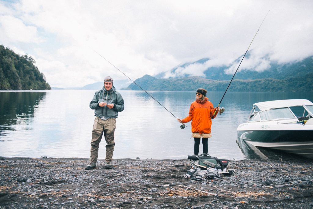 fishing quotes image of couple on shore holding fishing rods next to a boat. Mountains in the distance and cloudy sky