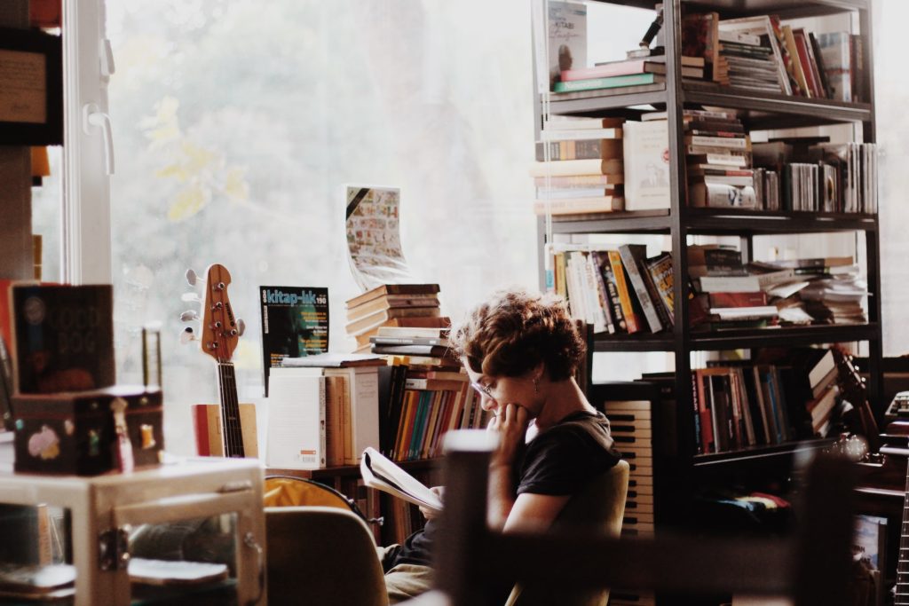 woman sitting in office area near window with stacks of books all around; reading hobby to practice patience