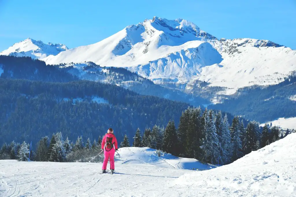 woman skier in pink snowsuit facing downhill looking at snow capped mountain in background