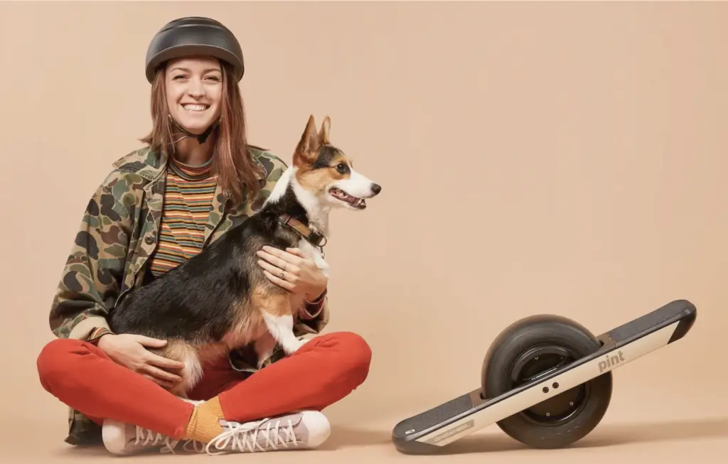 woman wearing a helmet, holding a dog with pint onewheel sitting next to her againts plain brown background