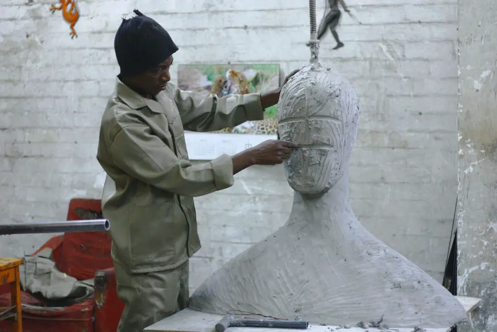 black man in hat sculpting a large bust indoors