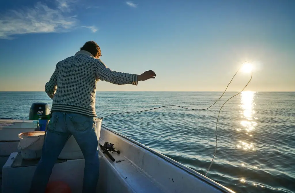 man magnet fishing from boat at sunrise, throwing rope out to the water, sun reflecting on water